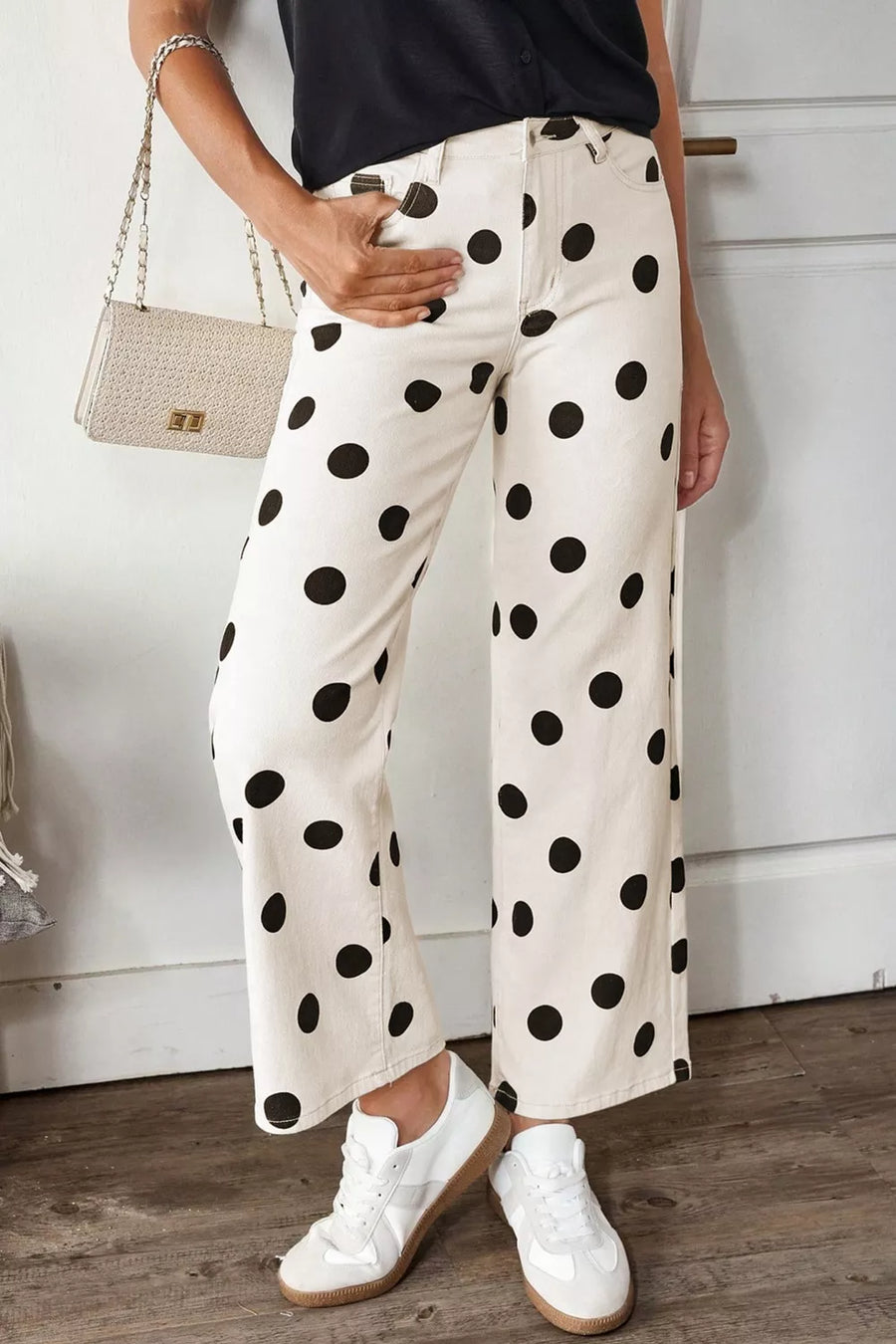 Person wearing white pants with black polka dots, white sneakers, and a black top, holding a beige handbag.