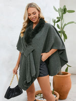 Woman wearing a green poncho with a fur collar, standing next to a potted plant.