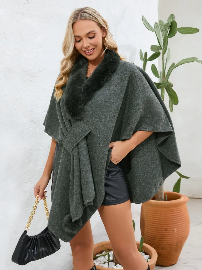 Woman wearing a green poncho with a fur collar, standing next to a potted plant.