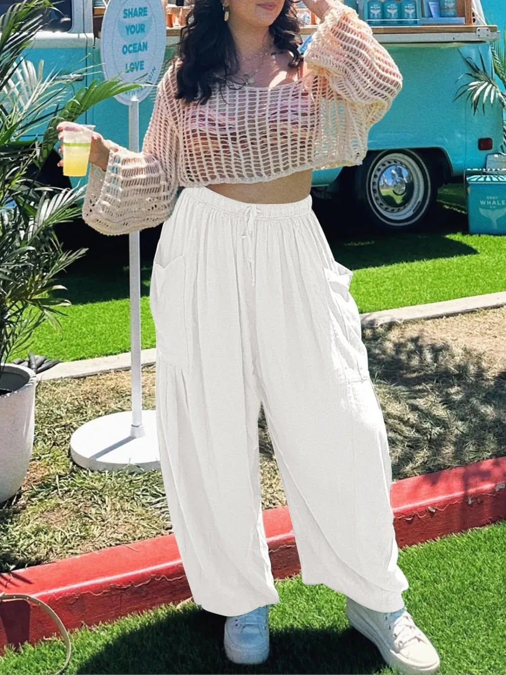 Person wearing a sheer top and white pants standing outdoors with a vehicle and plants in the background.