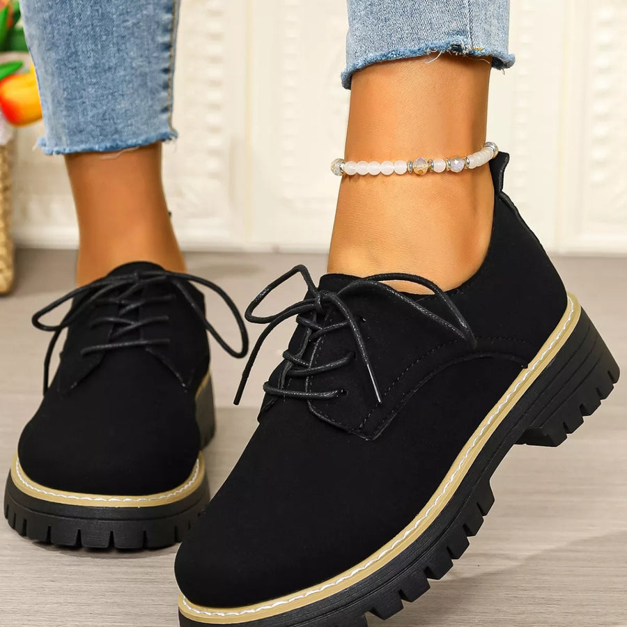 Black lace-up shoes with thick soles worn by a person, with a blurred indoor background.