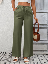 Person wearing green wide-leg pants with a white top and sandals, holding a brown handbag against a wooden wall.