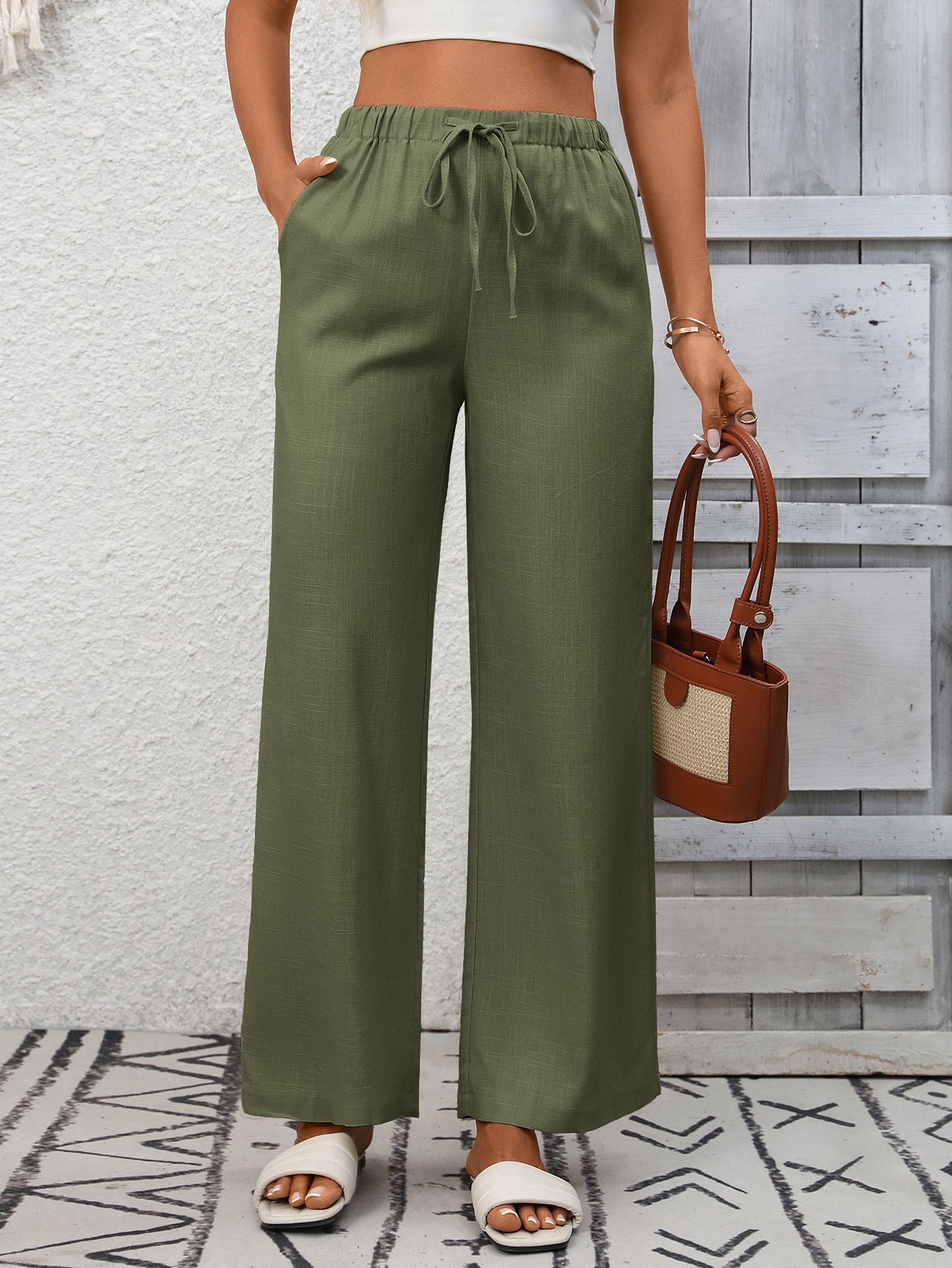 Person wearing green wide-leg pants with a white top and sandals, holding a brown handbag against a wooden wall.