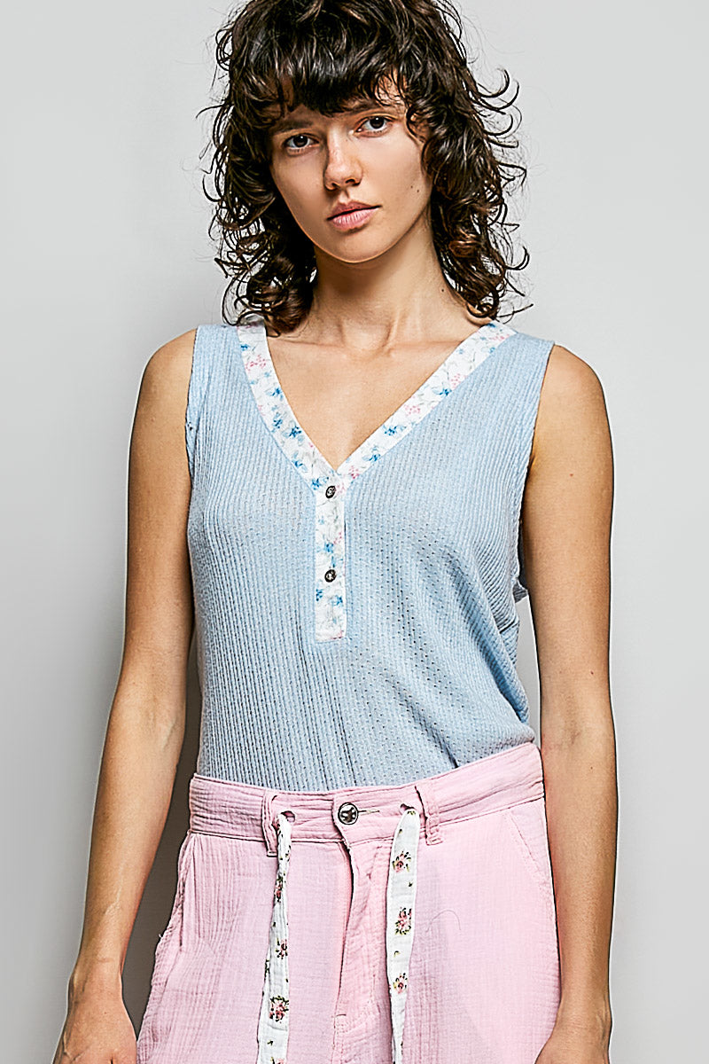 Woman wearing a light blue sleeveless top with floral trim and pink pants against a gray background