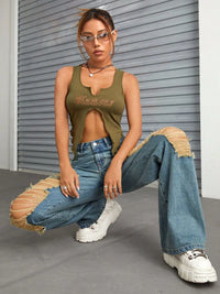 Person wearing a green tank top and blue jeans with ripped knees, squatting in front of a gray wall.