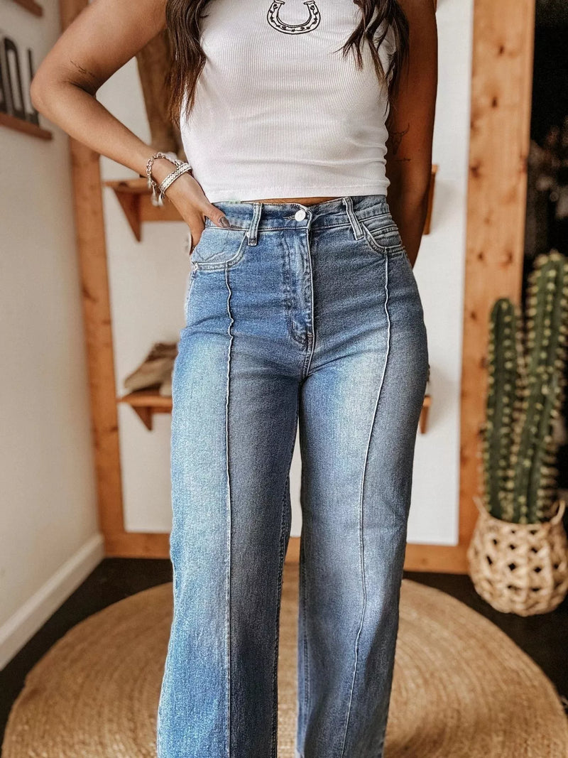 Person wearing a white tank top and blue jeans standing indoors with a wooden wall and cactus in the background.