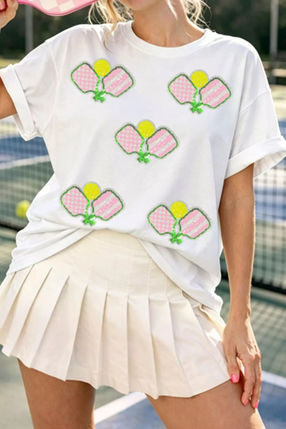 Person wearing a white t-shirt with colorful graphics and a beige skirt on a tennis court.