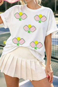 Person wearing a white t-shirt with colorful graphics and a beige skirt on a tennis court.