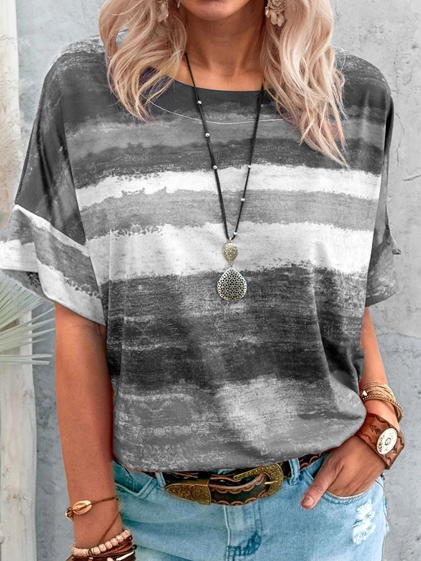 Person wearing a gray and white striped shirt with a necklace, standing against a concrete wall.
