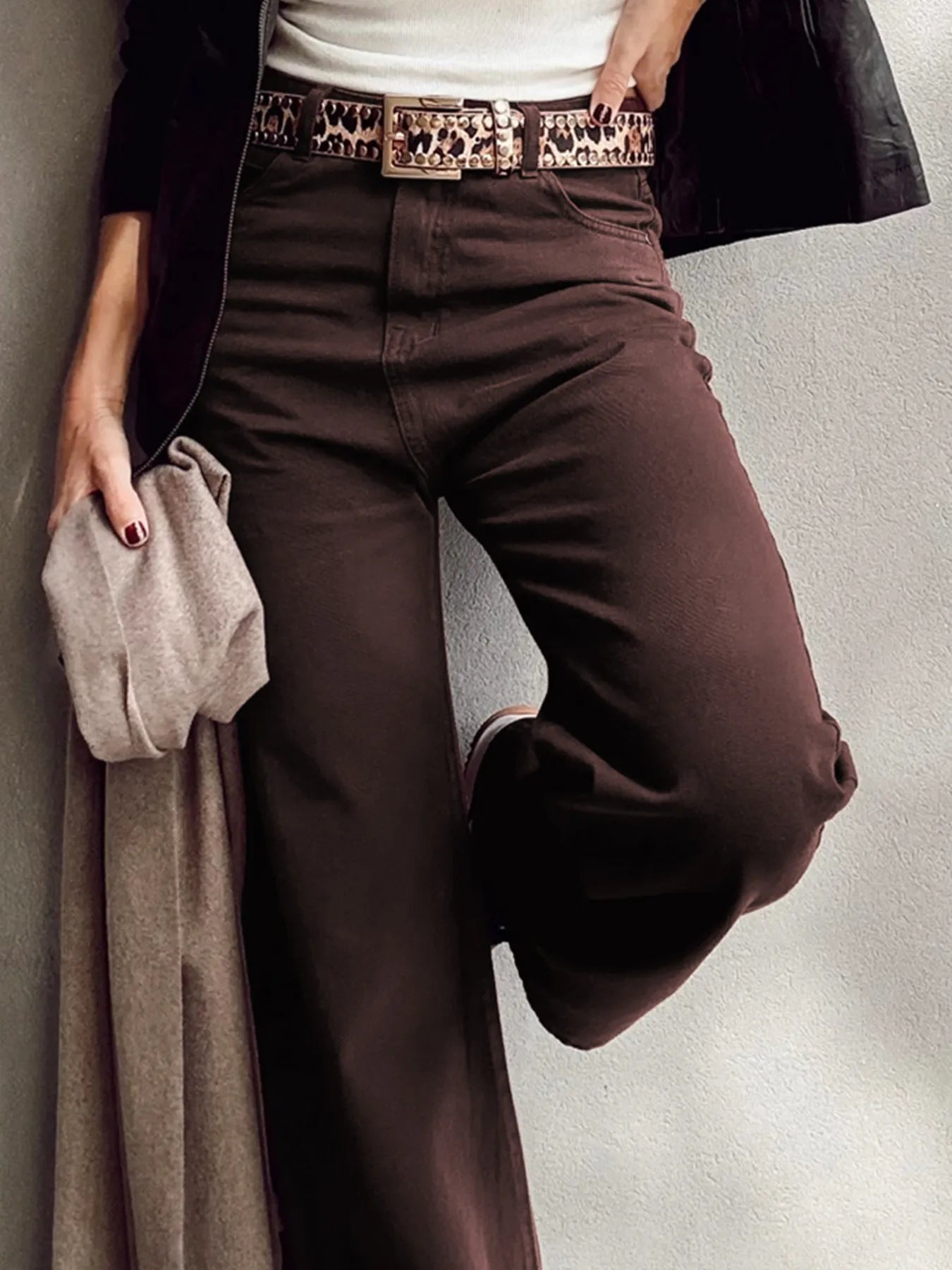 Person wearing brown pants with a patterned belt, holding a coat against a neutral background