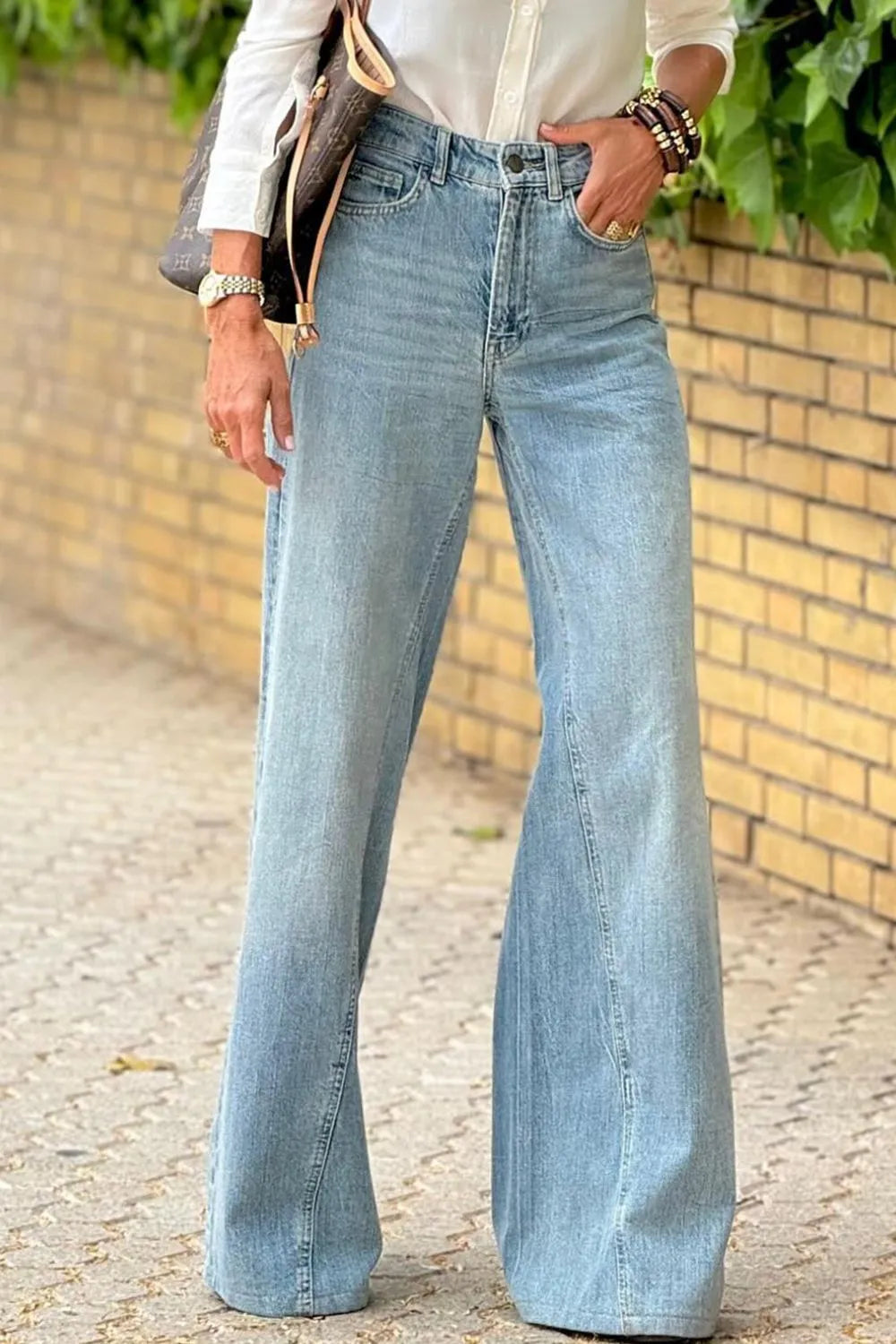 Person wearing light blue flared jeans standing on a cobblestone street with a brick wall in the background.