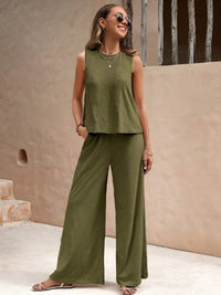 Woman wearing a green sleeveless top and wide-leg pants standing against a beige wall.