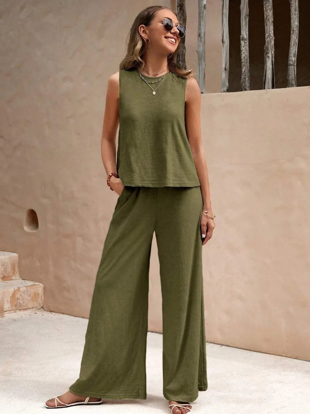 Woman wearing a green sleeveless top and wide-leg pants standing against a beige wall.