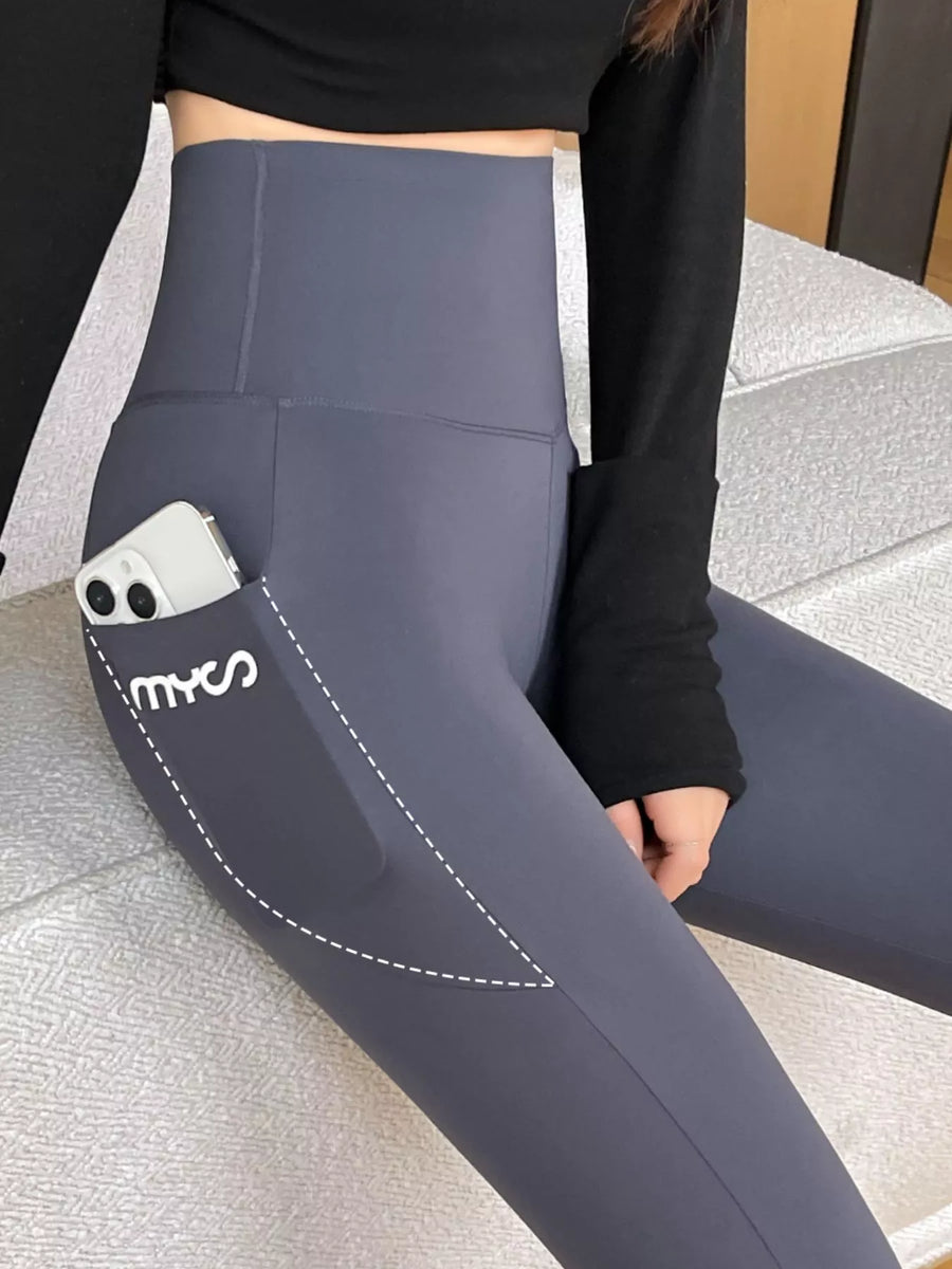 A close-up of a person wearing high-waisted slate gray leggings with a side pocket holding a white smartphone. The leggings have a white dashed line graphic and the brand name 'myos' printed on the pocket.
