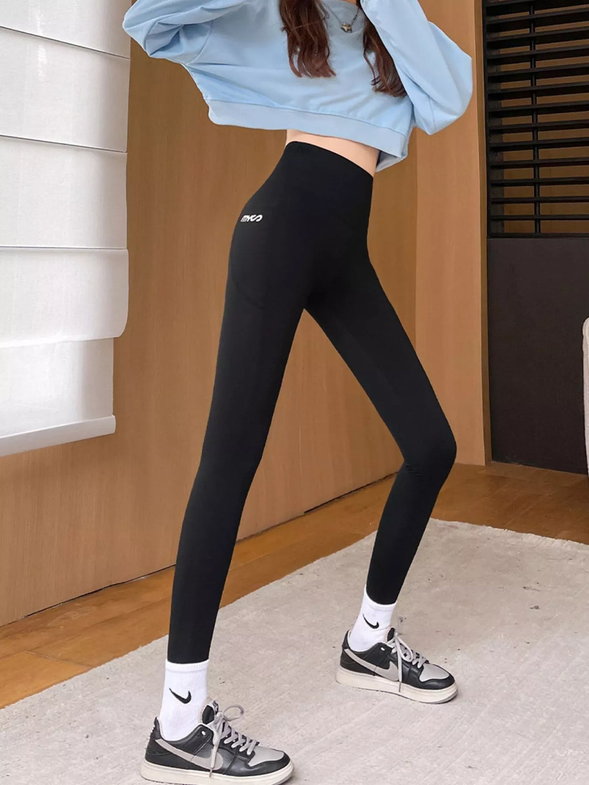 A person modeling black high-waisted leggings with a small white logo on the upper thigh, paired with white socks and black-and-white sneakers in an indoor room setting.