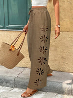Person wearing beige pants with floral cutouts, holding a brown bag against a stone wall.
