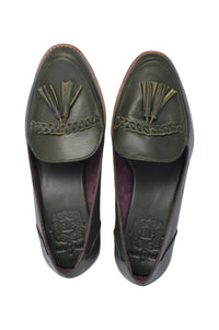 A pair of black leather loafers with tassels on the upper part, placed on a wicker surface. A person is in the process of fastening the shoes.