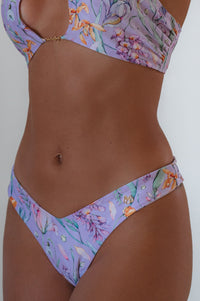 A close-up of a person wearing a brown Capri bikini bottom with a midrise waist and a dipped V front and back design, adorned with a gold signature charm.