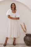 A woman wearing a white midi dress with puff sleeves and a square neckline, paired with black strappy sandals.