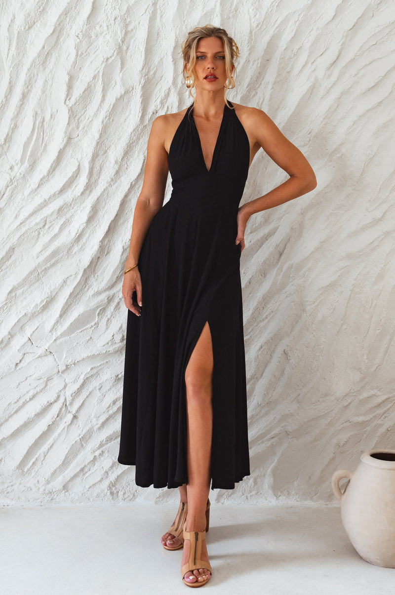 A woman wearing a black halter neck maxi dress with a fitted bodice and floaty skirt, featuring a front slit.
