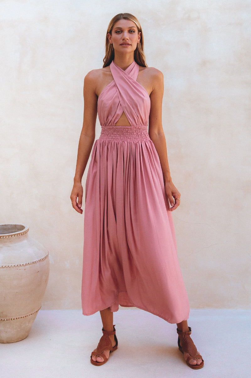 A woman wearing a pink halter neck, sleeveless, midi dress with elasticated waist and multi-way neck ties.