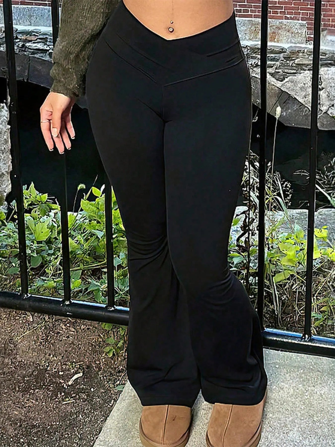 Person wearing black pants standing outdoors with a metal fence and greenery in the background