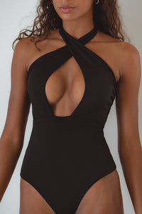 A woman lying on a striped towel wearing a brown one-piece swimsuit with a halter neck and cross-over design.