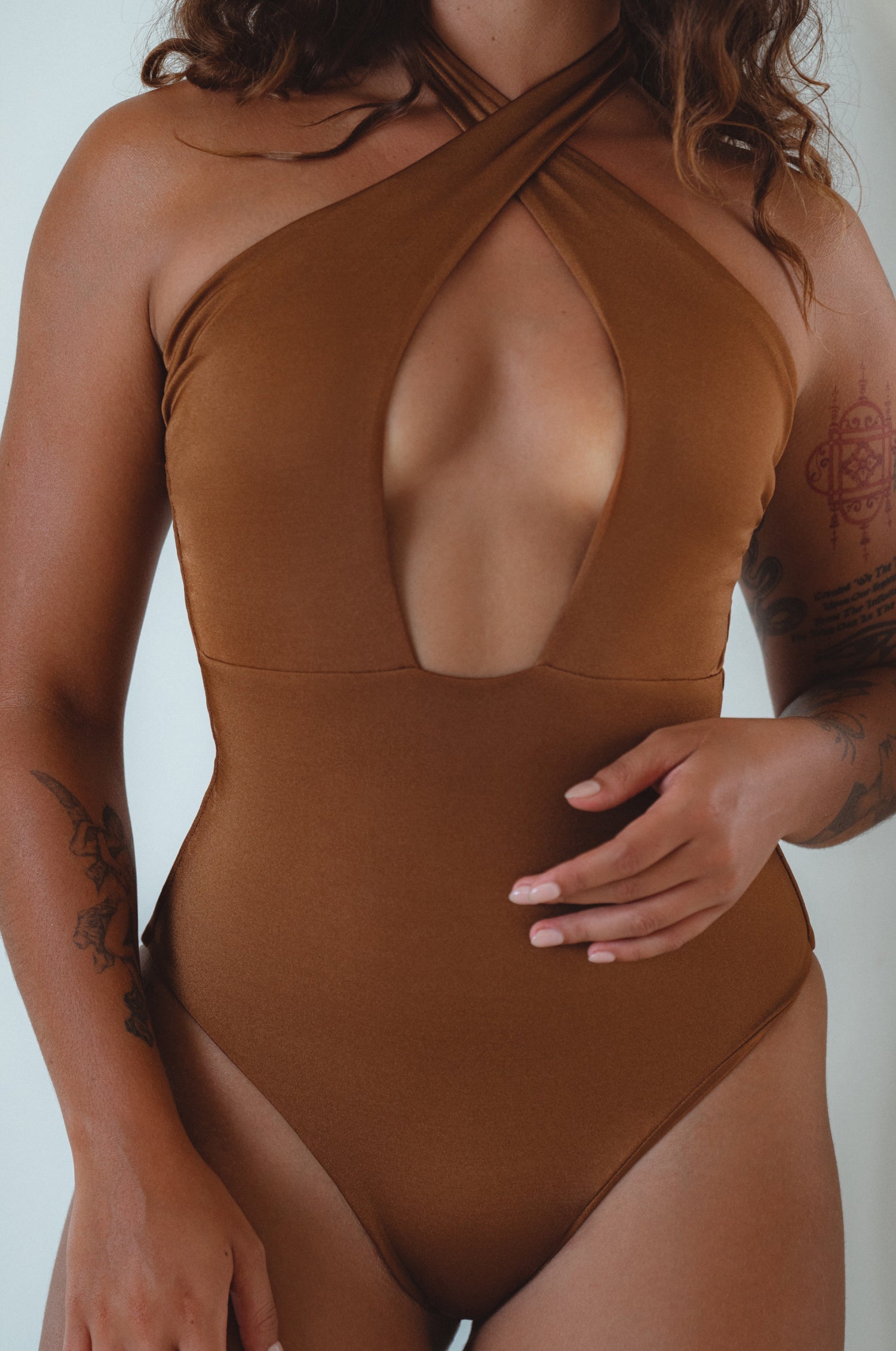 A woman lying on a striped towel wearing a brown one-piece swimsuit with a halter neck and cross-over design.