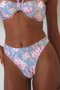A close-up of high-waisted red bikini bottoms with a bow detail at the front and a small branded charm on the side, modeled by a person.