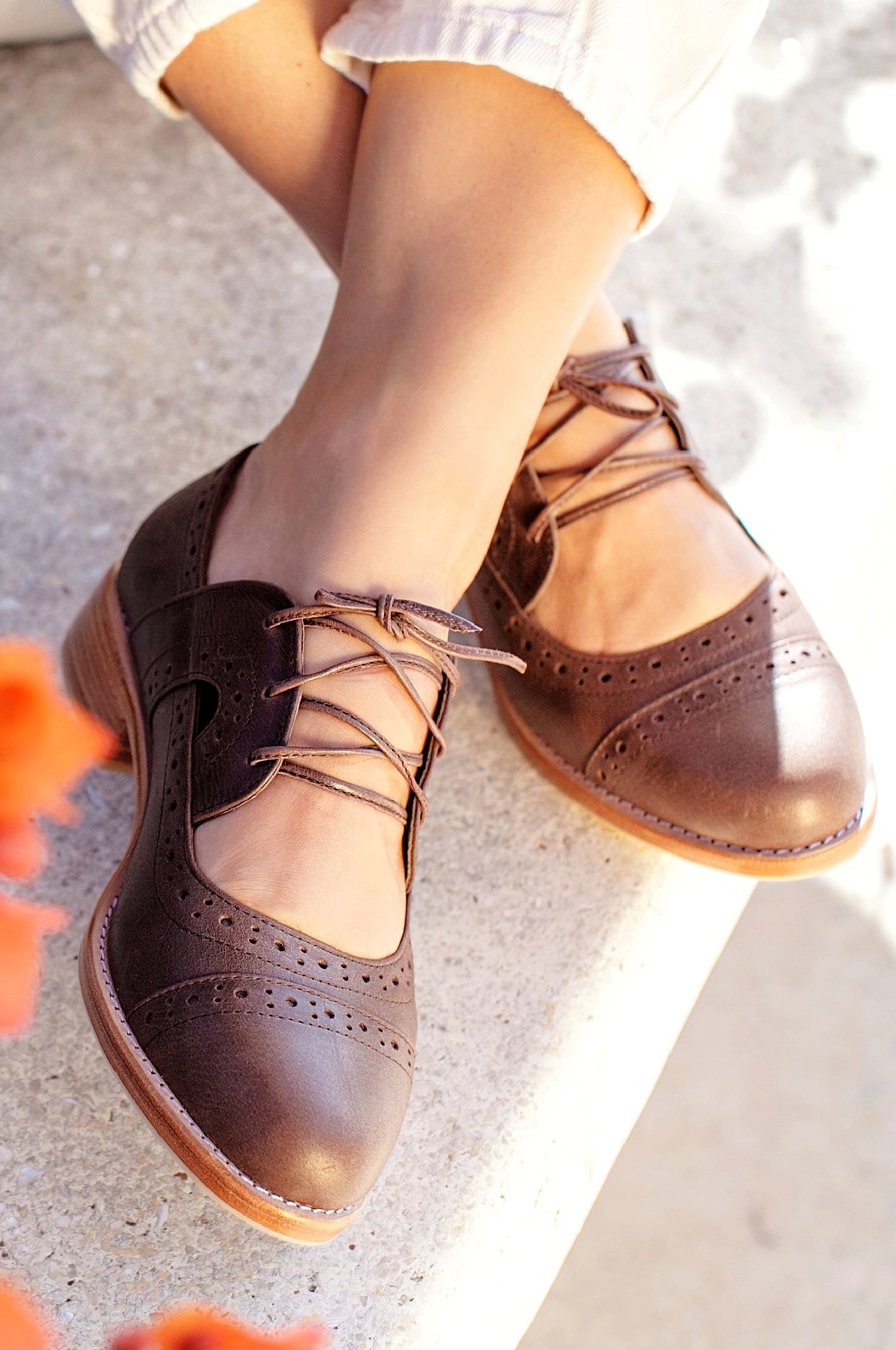 A pair of brown leather oxford shoes with lace-up closure, displayed on a person's feet against an outdoor setting.