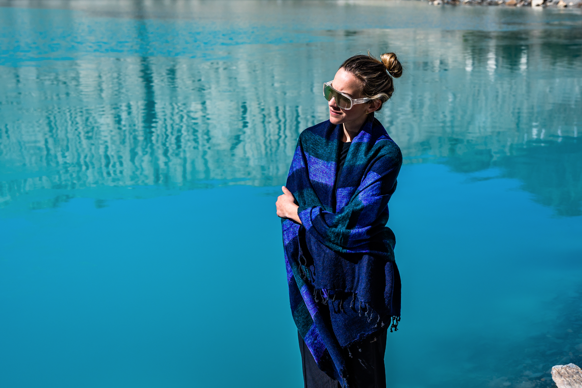 Woman standing by a body of water wearing a blue and black plaid shawl.