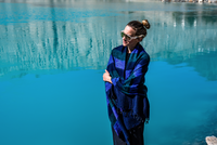 Woman standing by a body of water wearing a blue and black plaid shawl.