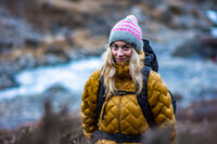 Woman in yellow puffer jacket and colorful knit hat standing in a natural setting with water in the background