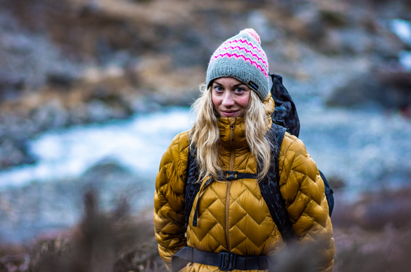 Woman in yellow puffer jacket and colorful knit hat standing in a natural setting with water in the background