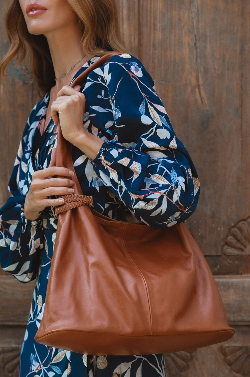 A woman wearing a floral dress carrying a brown leather tote bag with a shoulder strap.