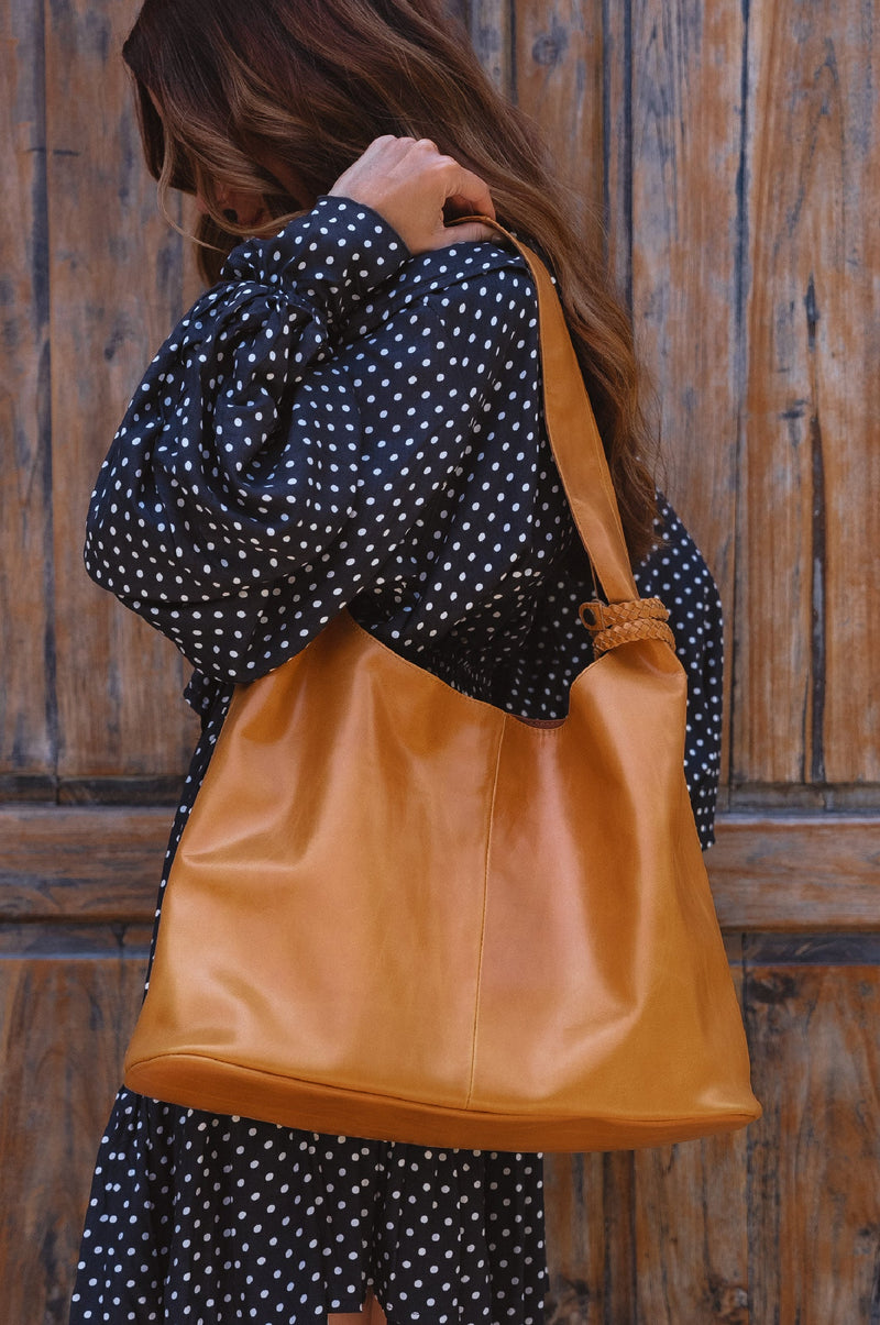 A person wearing a polka dot dress is holding a brown slouchy leather tote bag over their shoulder.