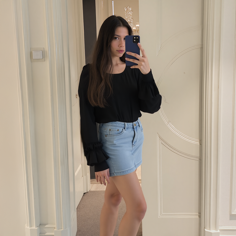 Woman taking a mirror selfie wearing a black top and denim skirt in an indoor setting.