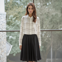 Woman wearing a checkered shirt and black skirt standing outdoors with trees in the background
