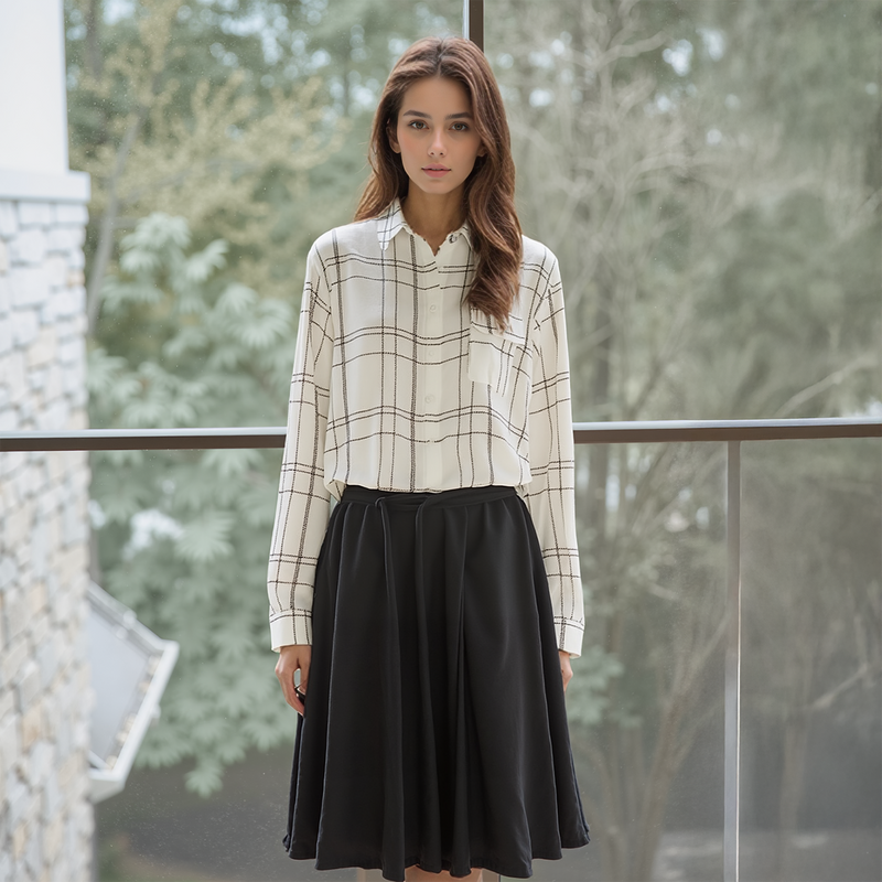 Woman wearing a checkered shirt and black skirt standing outdoors with trees in the background