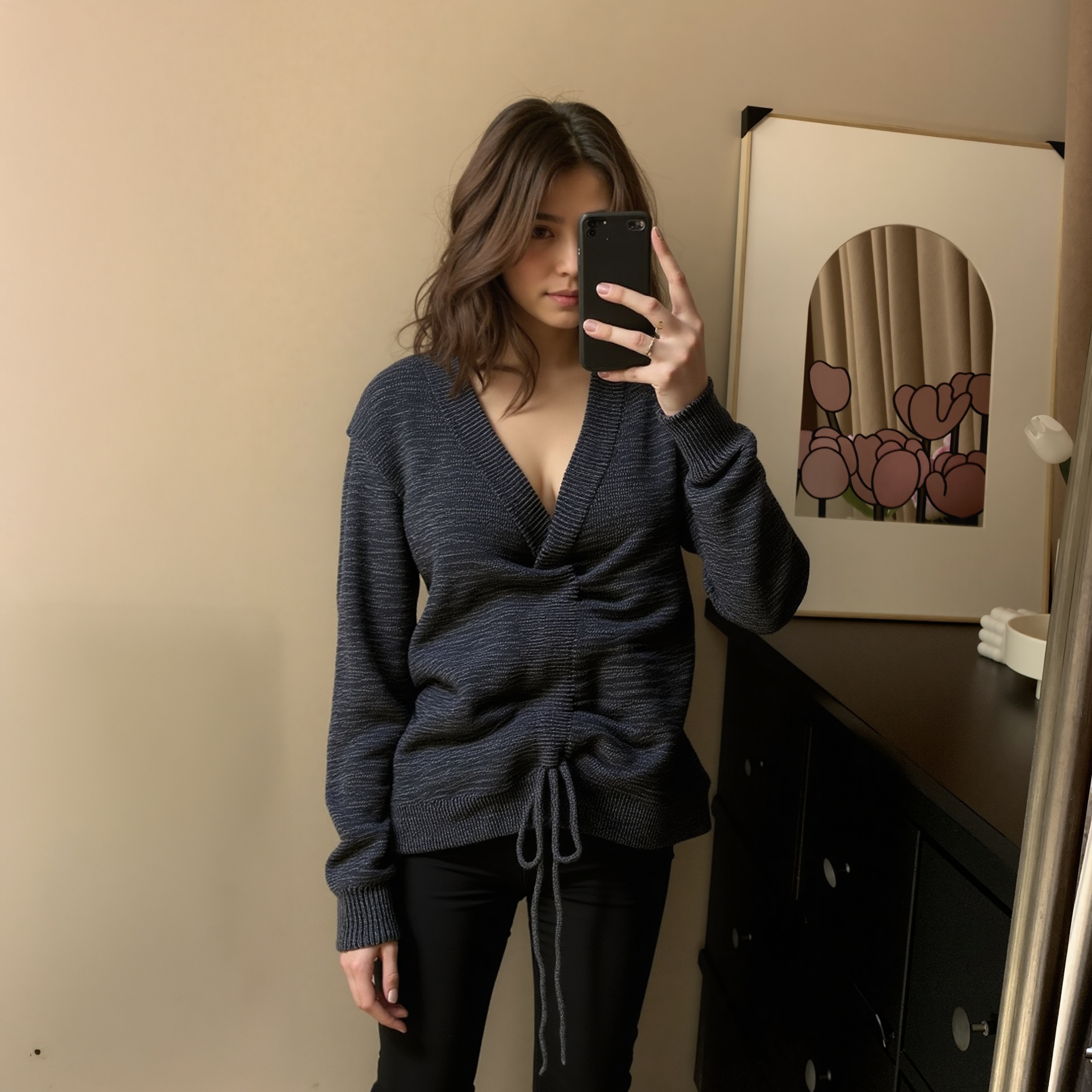 Woman taking a mirror selfie wearing a dark blue sweater in a room with a beige wall and a framed picture.