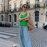 Woman in green top and light blue pants standing on a city street with classical architecture.