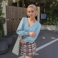 Woman in a light blue top and plaid skirt standing outdoors with a wooden fence and plants in the background.