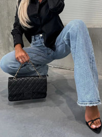 Person wearing a black jacket, blue jeans, and black sandals, holding a black quilted handbag.