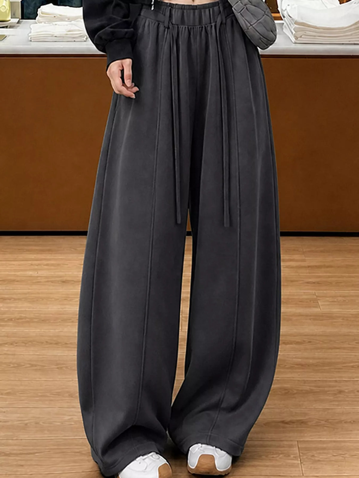 A person wearing very wide-leg dark charcoal gray pants with an elastic waistband and drawstrings, paired with a black top and white sneakers, standing on a wooden floor.