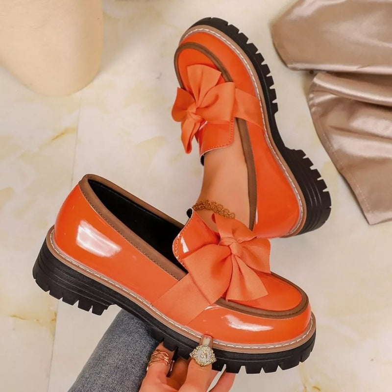 A pair of bright orange Mary Jane-style platform shoes with oversized satin bows and chunky lug soles, shown on a light marble-like surface with a hand holding one shoe.