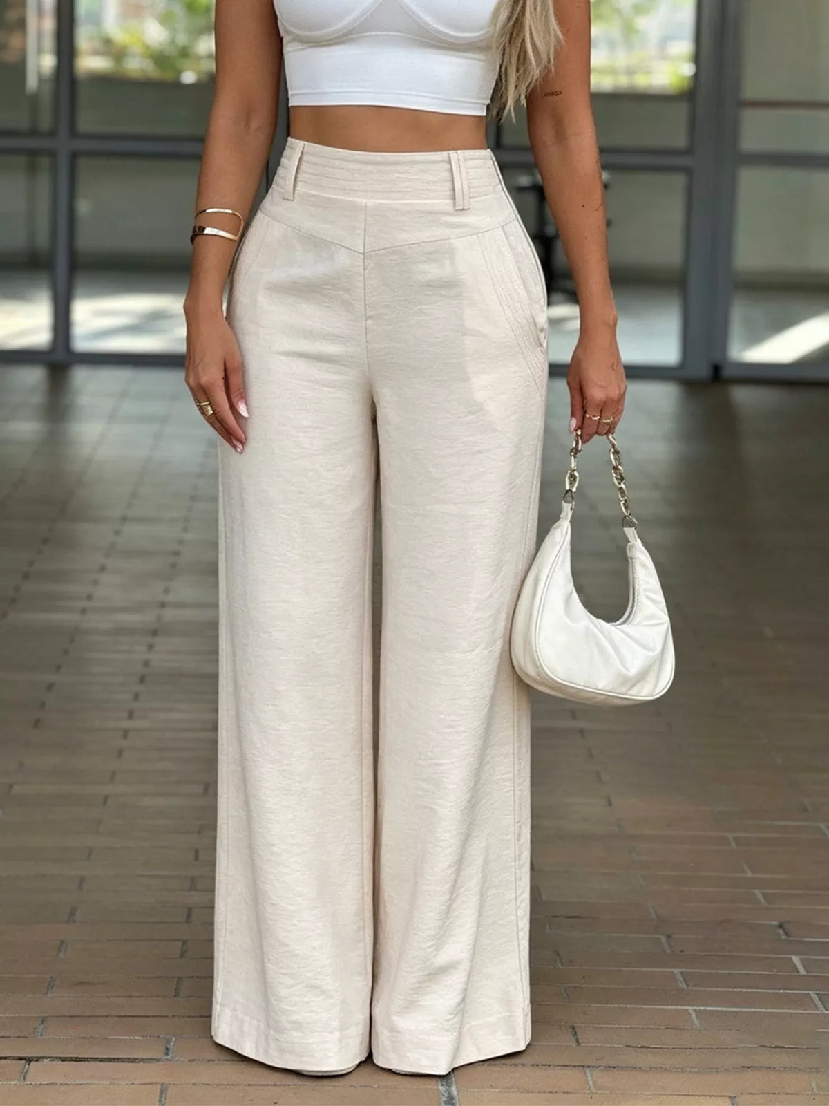 A woman wearing high-waisted, wide-leg cream trousers and a white crop top, holding a white crescent-shaped shoulder bag while standing on a tiled floor.
