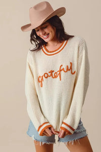 Woman wearing a cream sweater with 'grateful' text, denim shorts, and a beige hat on a beige background