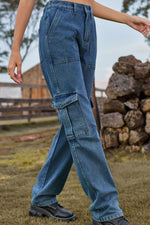 Person wearing blue jeans standing outdoors with a wooden fence and stacked logs in the background