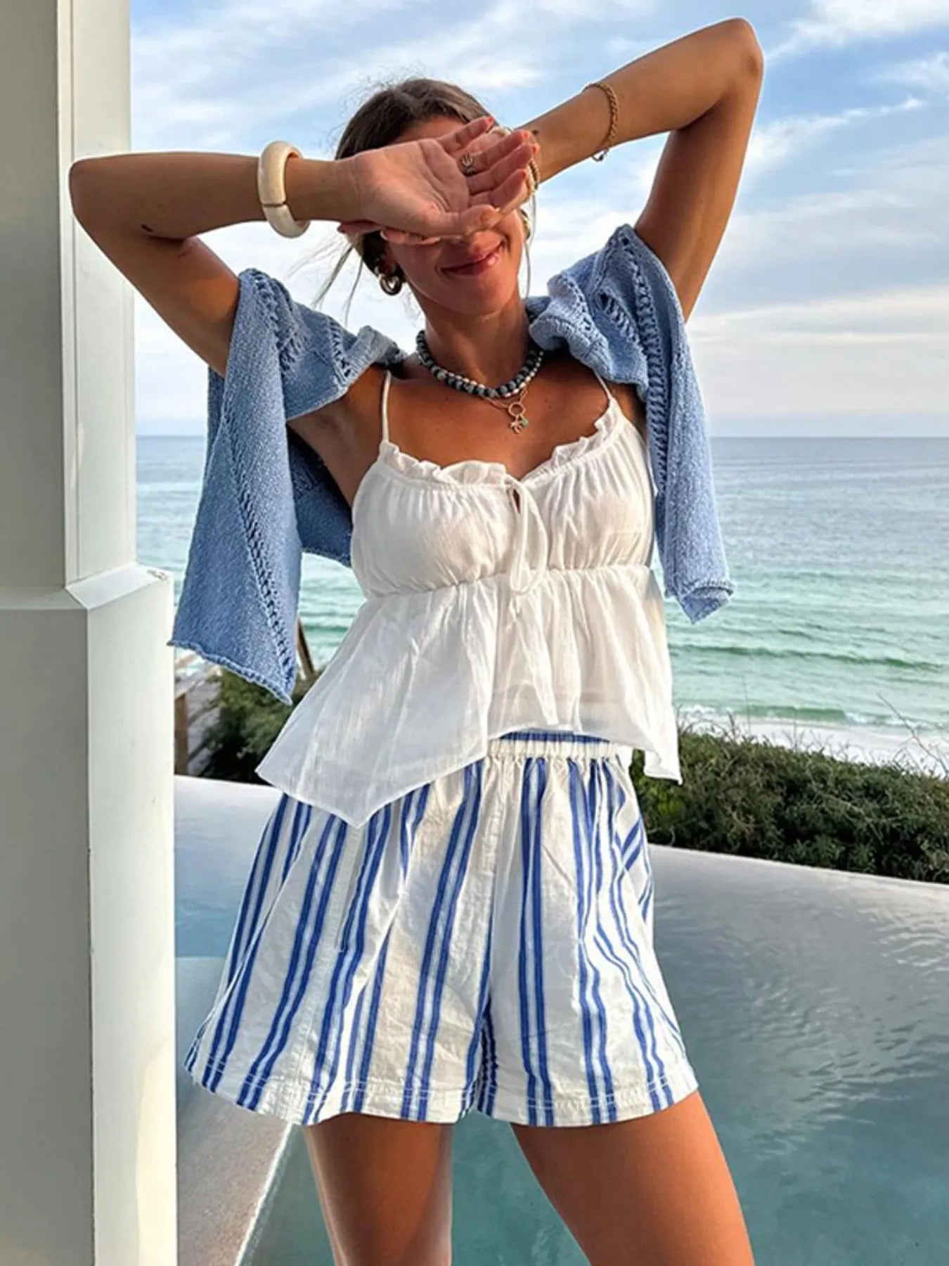 Woman in a white top and blue striped shorts standing by a pool with ocean view
