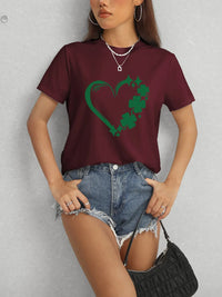 Person wearing a maroon t-shirt with a green heart design and denim shorts against a gray background